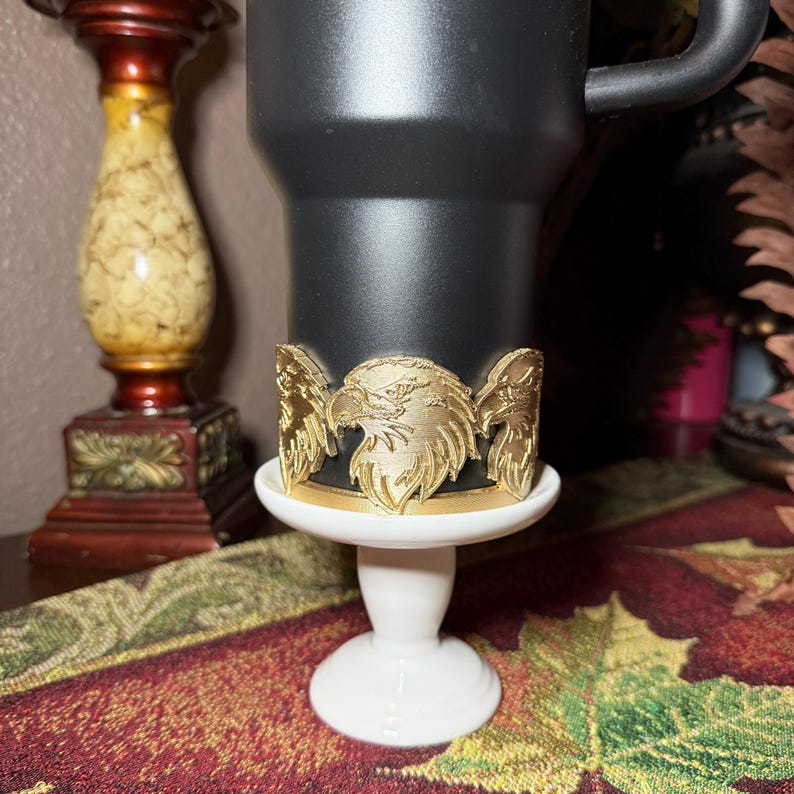 May include: A black tumbler with a gold-colored eagle emblem. The emblem is a detailed depiction of an eagle's head and wings. The tumbler sits on a white pedestal, and the background includes a decorative candle holder and a patterned tablecloth.