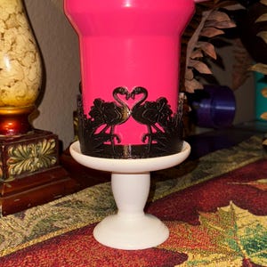May include: A bright pink tumbler featuring a black wrap-around cutout design of two flamingos facing each other with their necks forming a heart shape, set upon a white pedestal base with a smooth, rounded stem and a circular footed bottom.