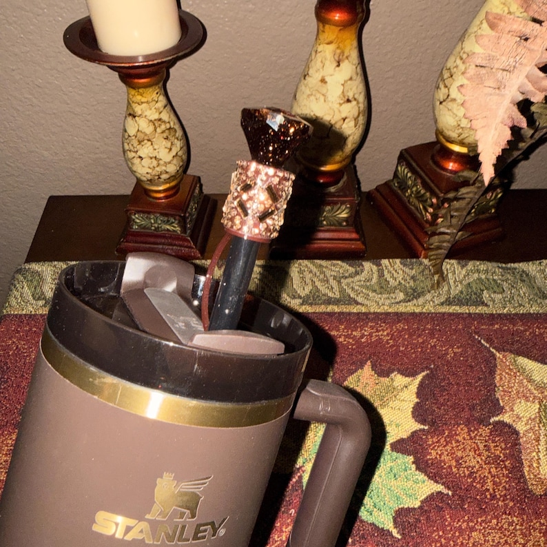 May include: A brown Stanley travel mug with a gold band and handle. A decorative bottle stopper with a rose gold and brown crystal design is inserted into the mug. The mug is on a table with decorative items.