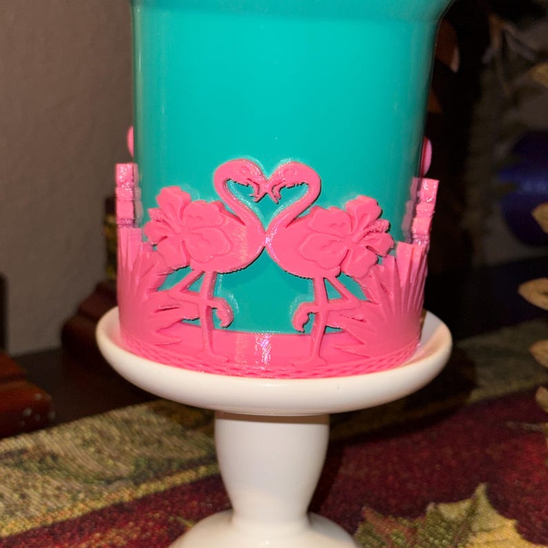 May include: A teal cylindrical container featuring a bright pink wrap-around overlay. The pink design displays two flamingos facing each other with their necks forming a heart shape, surrounded by hibiscus flowers and tropical palm leaves.