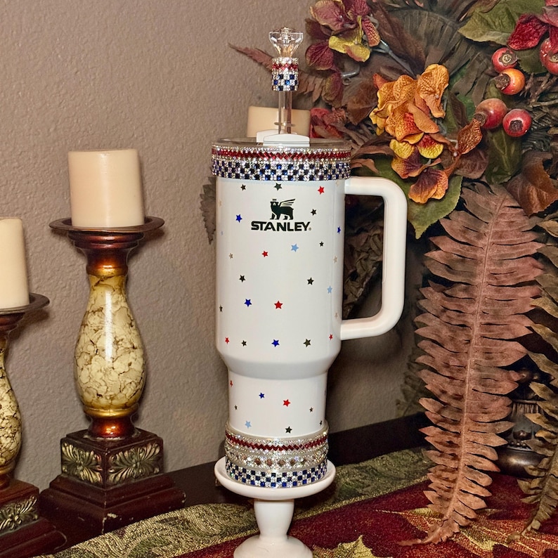 May include: A white Stanley travel mug with a handle, decorated with red and blue stars. The mug has a decorative rim and is mounted on a white stand. The Stanley logo is visible. A clear, decorative topper is on the lid.