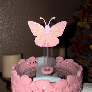 May include: A pink butterfly-shaped decorative object with yellow accents, mounted on a clear tube. The base is a light blue and pink floral design. The object is likely a cake topper or decorative item.