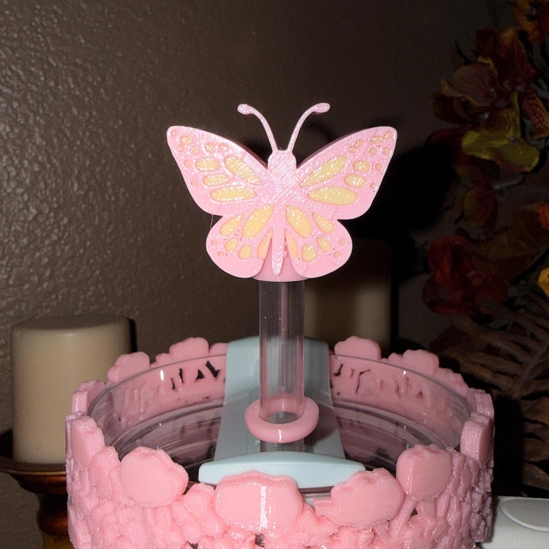 May include: A pink butterfly-shaped decorative object with yellow accents, mounted on a clear tube. The base is a light blue and pink floral design. The object is likely a cake topper or decorative item.