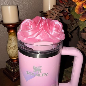 May include: A pink Stanley mug with a rose-shaped lid. The mug has a handle and the Stanley logo. The mug is decorated with a heart design. The lid is a light pink color and shaped like a rose.