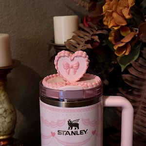 May include: A pink Stanley travel mug with a heart-shaped topper and bow design. The mug features a decorative pattern and the Stanley logo. The handle and lid are also pink. The mug is designed for beverages.