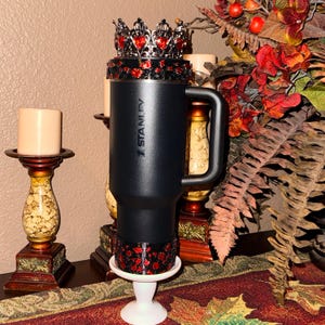 May include: A black Stanley travel mug with a decorative red and black crown and base. The mug has a handle and is displayed on a white pedestal. The background includes candles and fall foliage.