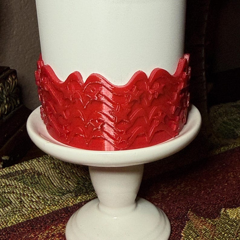 May include: A white cylindrical object sits on a white pedestal stand. A bright red band wraps around the base of the cylinder, featuring a repeating embossed pattern of five-pointed stars and wavy lines that create a textured, decorative border.