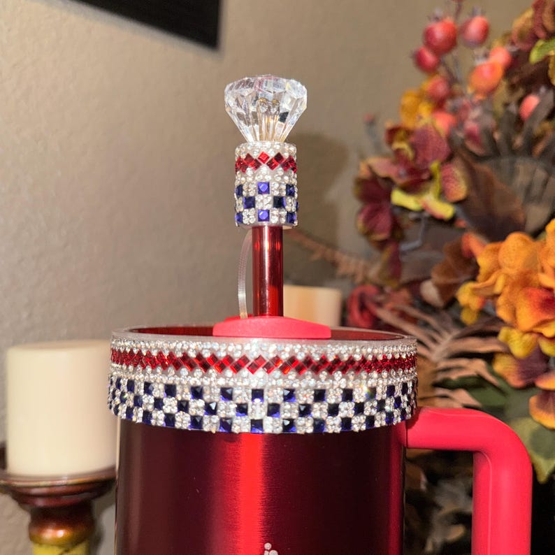 May include: A red tumbler with a decorative rim and a handle. The tumbler is adorned with red, white, and blue rhinestones. A clear, faceted gem tops the red straw. The design is reminiscent of a patriotic theme.