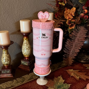 May include: A pink Stanley travel mug with a decorative cake design, a heart-shaped lid, and a handle. The mug features the Stanley logo and cherry illustrations. It is displayed on a white pedestal.