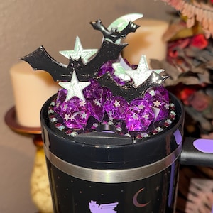 May include: Black travel mug with a purple and black bat and star design. The lid is decorated with purple gems, black bats, and white stars. The handle is purple.
