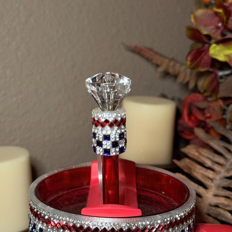 May include: A decorative bottle stopper with a large, clear, diamond-shaped top. The stopper's body is adorned with red, white, and blue rhinestones. The stopper is set in a red holder, and the rim of the base is decorated with rhinestones.