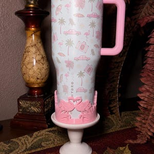 May include: A light blue stainless steel tumbler with a pink handle and lid, featuring a repeating pattern of pink flamingos, palm trees, cars, and suitcases. The word HYDROJUG is printed vertically. A pink flamingo-shaped base stand is attached.