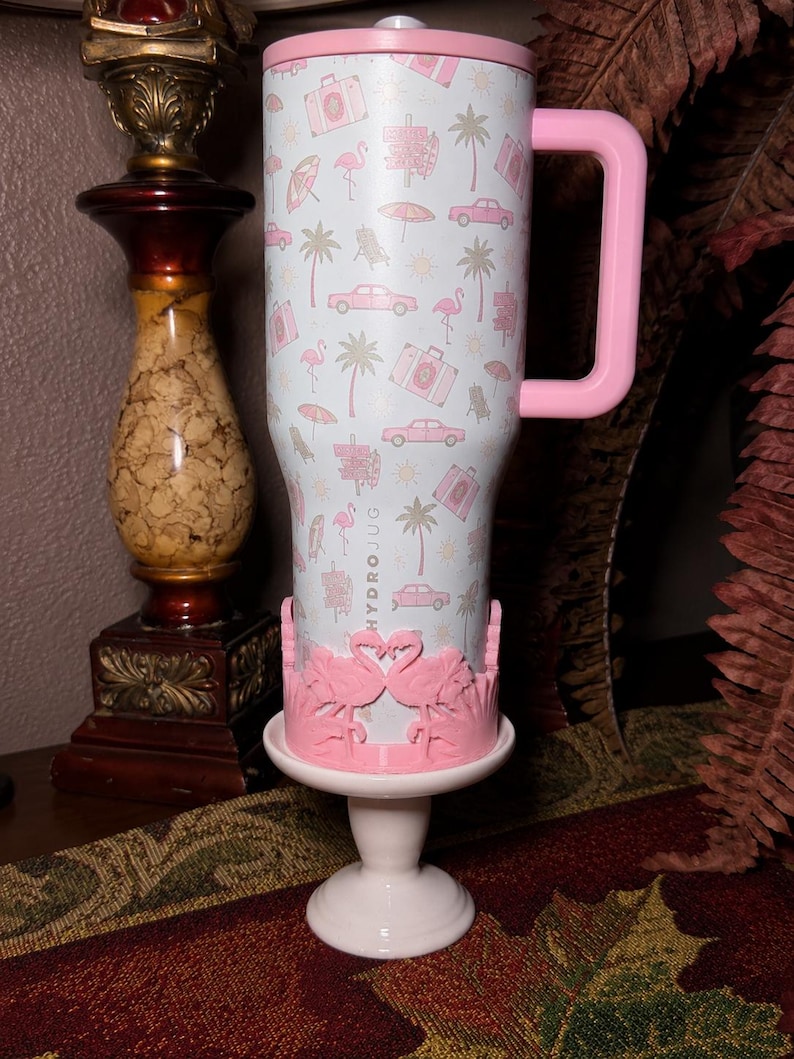 May include: A light blue stainless steel tumbler with a pink handle and lid, featuring a repeating pattern of pink flamingos, palm trees, cars, and suitcases. The word HYDROJUG is printed vertically. A pink flamingo-shaped base stand is attached.