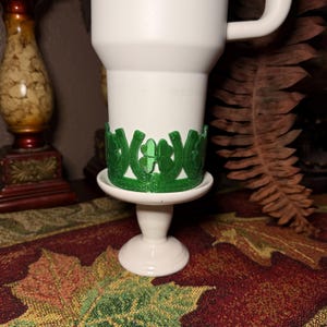 May include: A white travel mug with a green shamrock and horseshoe design. The mug sits on a white pedestal with a green shamrock and horseshoe design. The mug is on a table with a fall-themed tablecloth.
