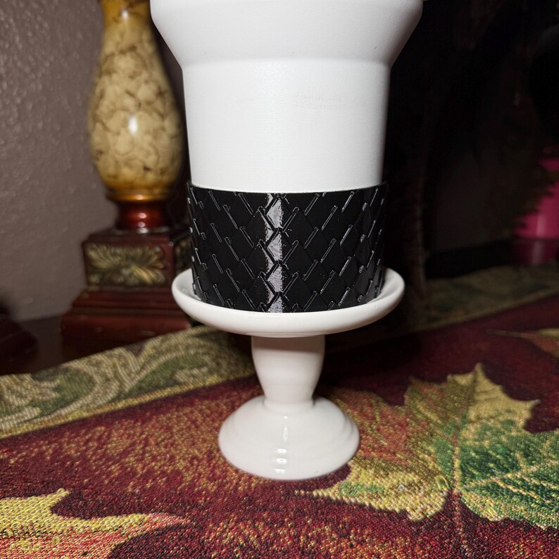 May include: A white ceramic candle holder with a black woven pattern band. The candle holder has a pedestal base and a wide, shallow dish to hold the candle. The design is simple and elegant, suitable for various home decor styles.