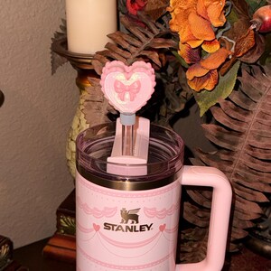 May include: A pink Stanley mug with a heart-shaped lid ornament. The mug has a decorative design and the Stanley logo. The heart-shaped ornament has a bow design. The mug has a handle.