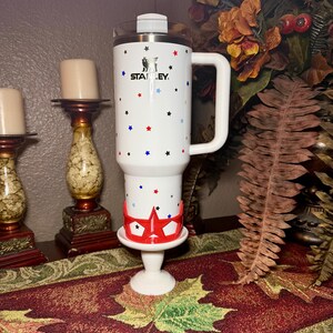 May include: White Stanley travel mug with a handle, decorated with red, blue, and black stars. The mug has a silver lid and the Stanley logo. It is placed on a white pedestal with a red star-shaped holder.
