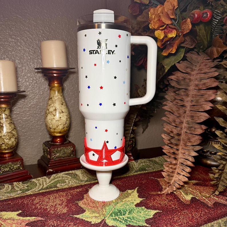 May include: White Stanley travel mug with a handle, decorated with red, blue, and black stars. The mug has a silver lid and the Stanley logo. It is placed on a white pedestal with a red star-shaped holder.