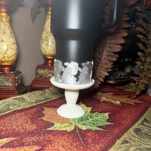 May include: A black tumbler with a decorative silver metal wrap featuring a cowgirl silhouette design. The tumbler sits on a white pedestal base, placed on a patterned red and green table runner. Decorative items are in the background.