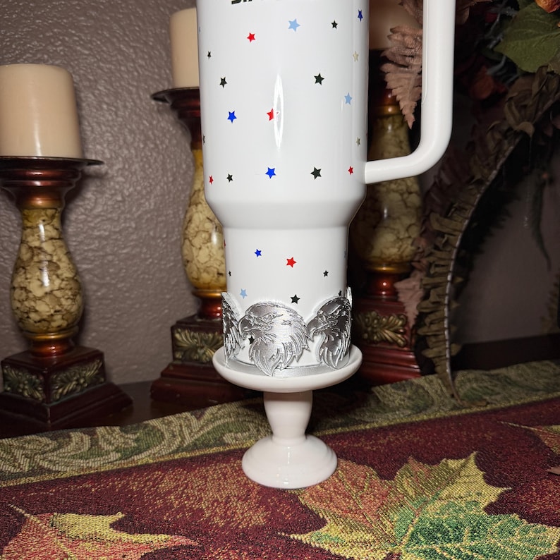 May include: White travel mug with a handle, decorated with red, blue, and black stars. The mug sits on a white stand with a silver eagle design. The mug is a patriotic theme.