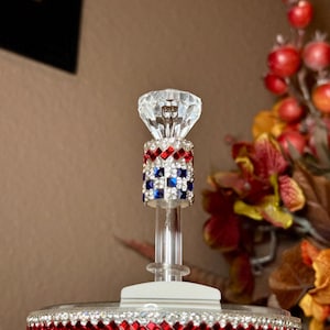 May include: A decorative bottle stopper with a clear, faceted crystal top. The stopper's stem is adorned with red, white, and blue rhinestones in a checkered pattern. The base is also decorated with red rhinestones. The background features fall foliage.