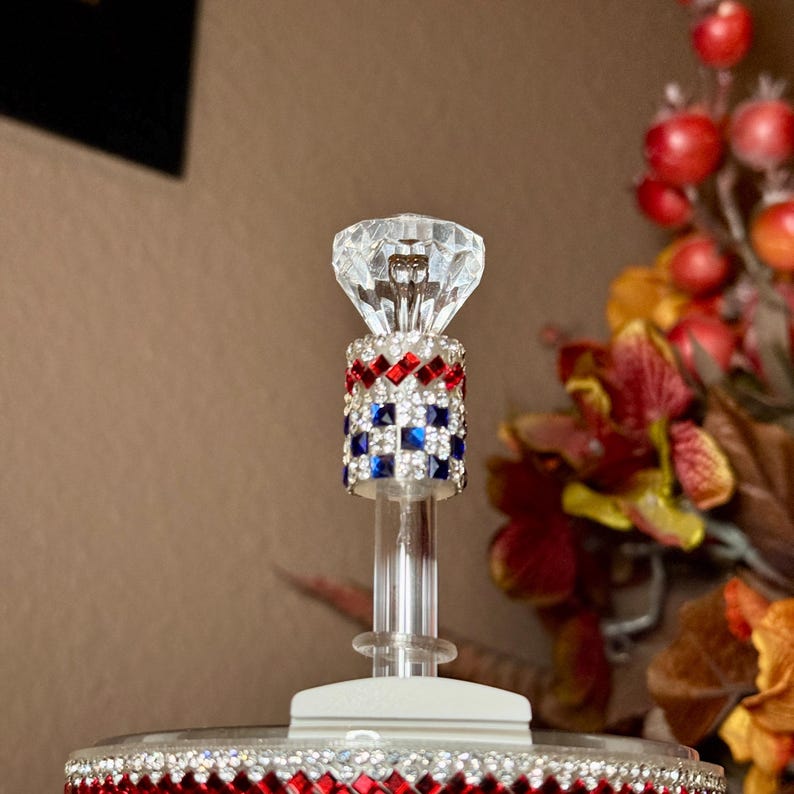 May include: A decorative bottle stopper with a clear, faceted crystal top. The stopper's stem is adorned with red, white, and blue rhinestones in a checkered pattern. The base is also decorated with red rhinestones. The background features fall foliage.