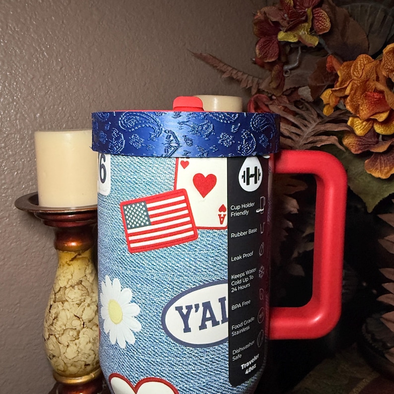 May include: A blue denim-patterned tumbler with a red handle and a blue paisley lid. The surface features stickers including an American flag, a heart-shaped playing card, a white daisy, and the text Y'ALL. A black label lists features like 40oz.