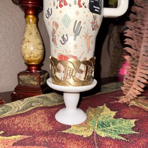 K&ouml;nnte beinhalten: Ein wei&szlig;er Reisebecher mit Cowboy-Motiv, einschlie&szlig;lich Stiefeln, Kakteen und Sternen. Der Becher wird von einem goldfarbenen Metallhalter auf einem wei&szlig;en Sockel gehalten. Der Becher ist mit "Traveler 40oz." beschriftet.