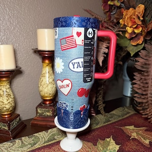 May include: A 40oz stainless steel tumbler with a denim-textured blue exterior featuring stickers of an American flag, a daisy, cherries, dice, and the text Y'ALL and DARLIN'. It has a red handle, a dark blue paisley lid, and a dark blue base.