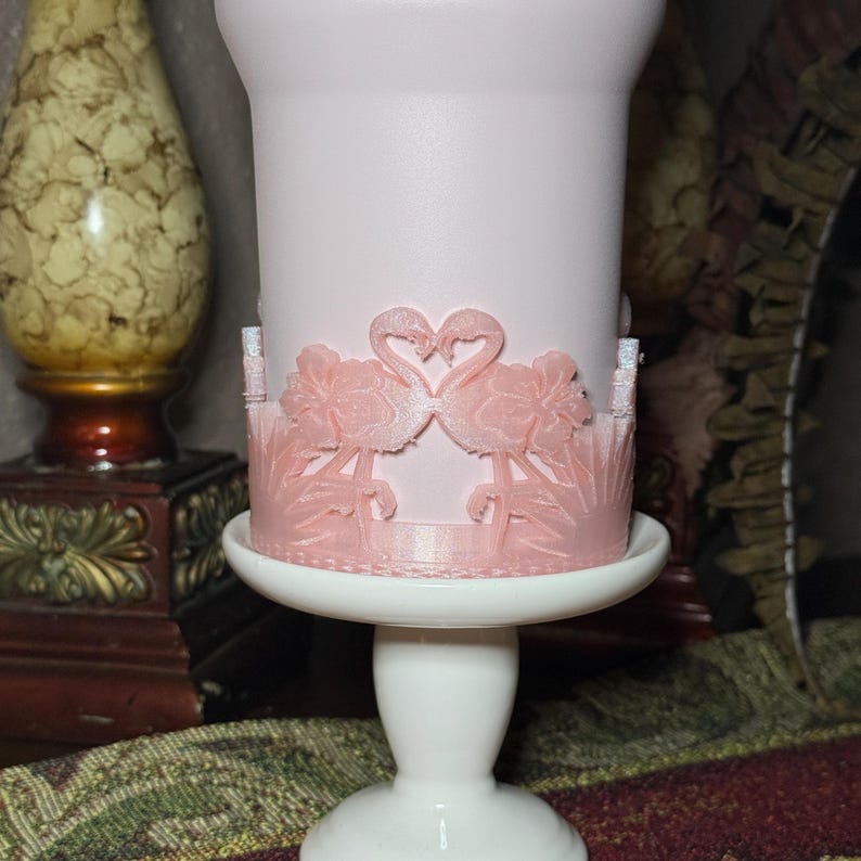 May include: A light pink cylindrical tumbler featuring a raised, textured wrap around the base. The wrap displays two flamingos facing each other with their necks forming a heart shape, surrounded by hibiscus flowers and tropical foliage.