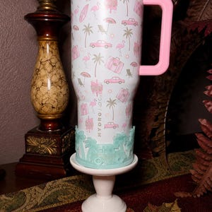 May include: A white stainless steel tumbler with a pink lid and handle, featuring a repeating pattern of pink flamingos, palm trees, vintage cars, suitcases, and motel signs. The word HYDROJUG is printed vertically. A light green silicone boot is attached.