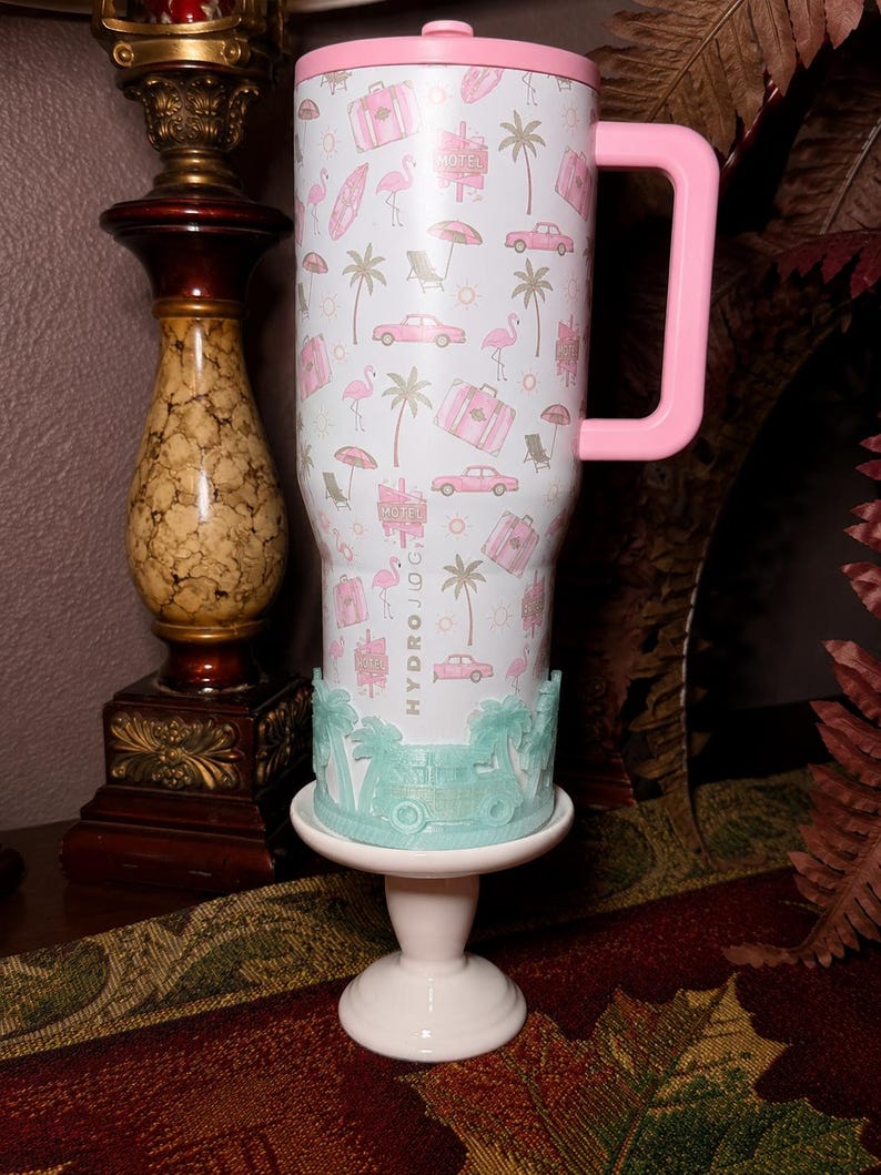 May include: A white stainless steel tumbler with a pink lid and handle, featuring a repeating pattern of pink flamingos, palm trees, vintage cars, suitcases, and motel signs. The word HYDROJUG is printed vertically. A light green silicone boot is attached.