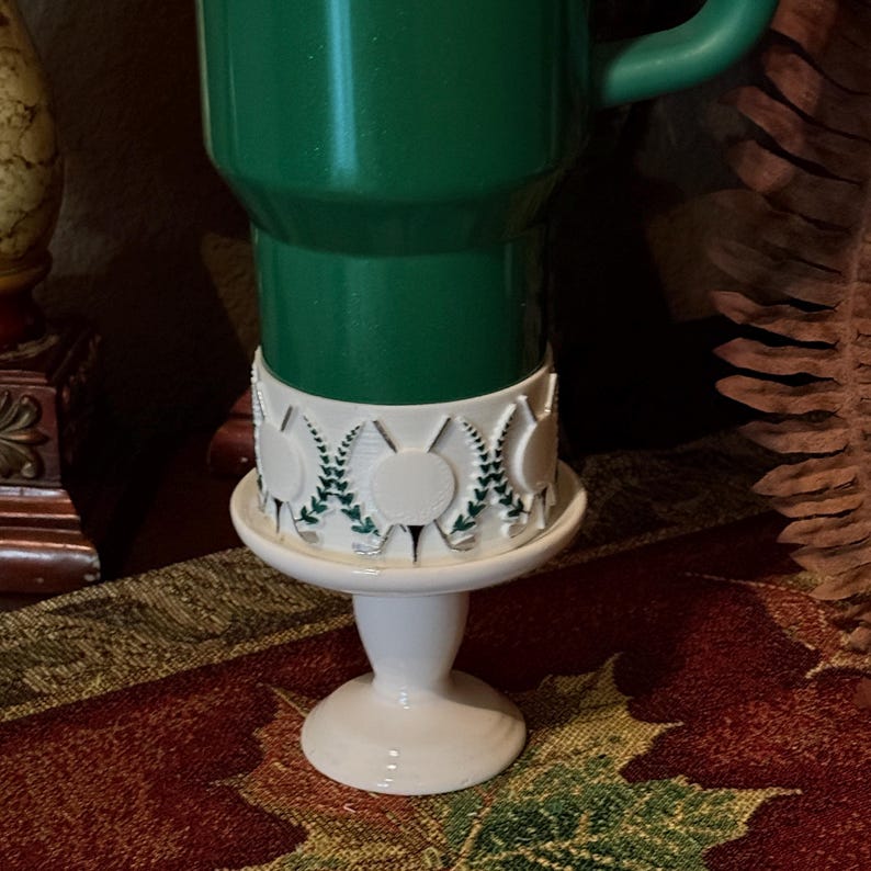 May include: A green mug sits atop a white pedestal with a decorative band featuring circular designs and green leaf accents. The pedestal has a round base and a small, round top. The mug is a solid, vibrant green.