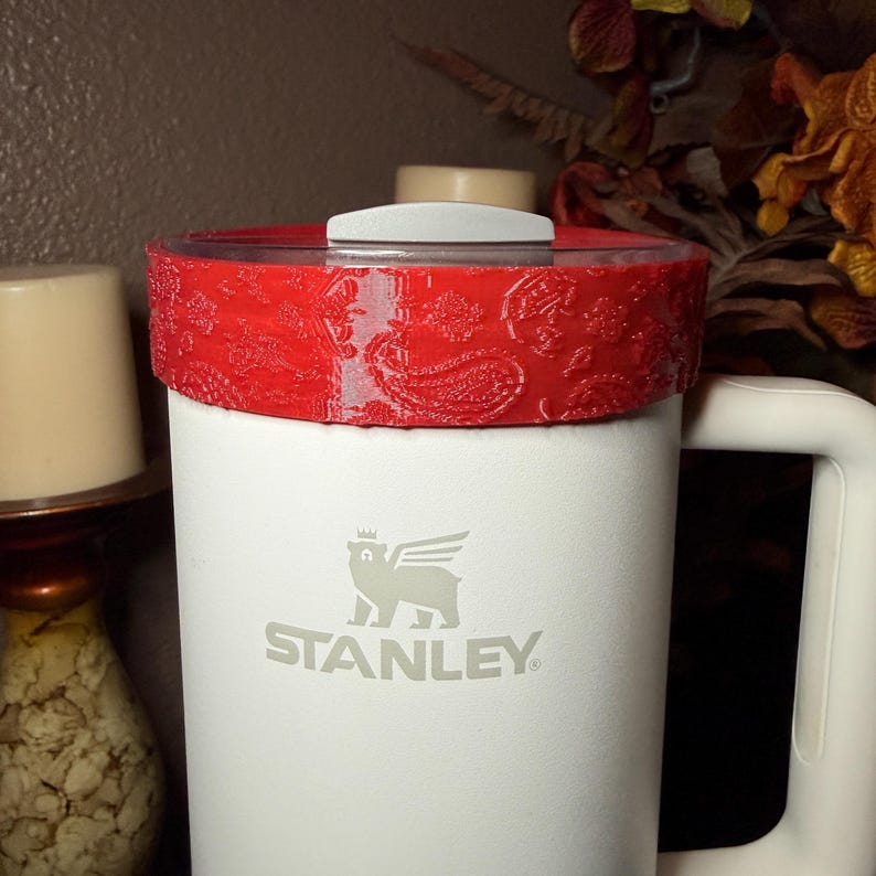 May include: A white Stanley tumbler featuring a handle and a red lid topper with a textured paisley pattern. The front of the tumbler displays the grey Stanley logo, which includes a winged bear icon positioned above the brand name text.
