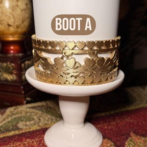 May include: A white pedestal candle holder with a gold-colored decorative band featuring heart and floral cutouts. The text "BOOT A" is on the candle holder. The candle holder is on a patterned surface.