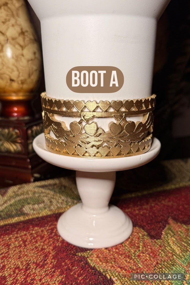 May include: A white pedestal candle holder with a gold-colored decorative band featuring heart and floral cutouts. The text "BOOT A" is on the candle holder. The candle holder is on a patterned surface.