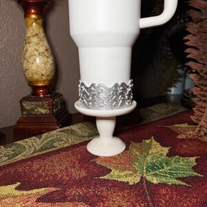 May include: A white tumbler with a handle, featuring a silver-colored band around the base with a repeating wave and star pattern. The tumbler is supported by a white pedestal stand, creating a raised display for the drinkware item.
