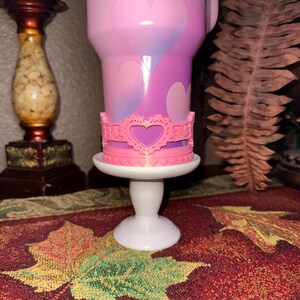 Puede incluir: Un vaso rosa y morado con diseños de corazones, adornado con una corona rosa en forma de corazón. El vaso se encuentra sobre un pedestal blanco, colocado sobre una superficie estampada con hojas de otoño.