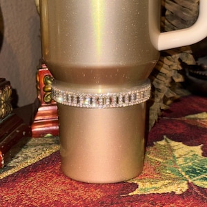 May include: A gold glitter tumbler with a rhinestone band around the middle. The tumbler has a white lid and handle.