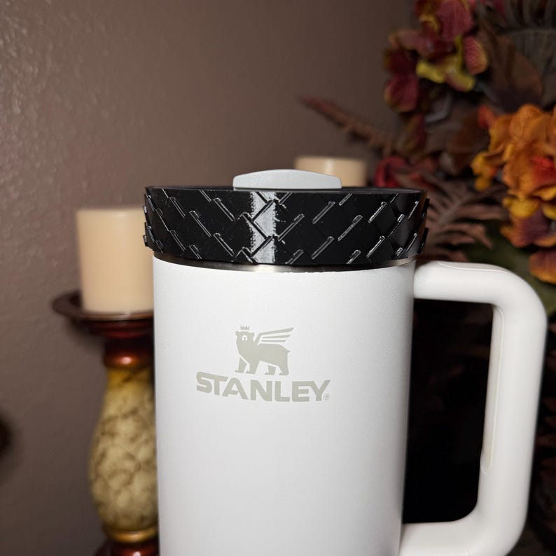 May include: White Stanley mug with a black textured lid and a white handle. The Stanley logo is printed in gray on the front of the mug. The mug is cylindrical and appears to be made of stainless steel.
