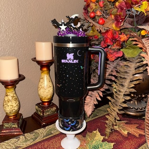 May include: Black Stanley travel mug with a handle, decorated with bats, stars, and a crescent moon. The mug has a decorative topper with bats and stars. The mug is on a white pedestal. The Stanley logo is visible.