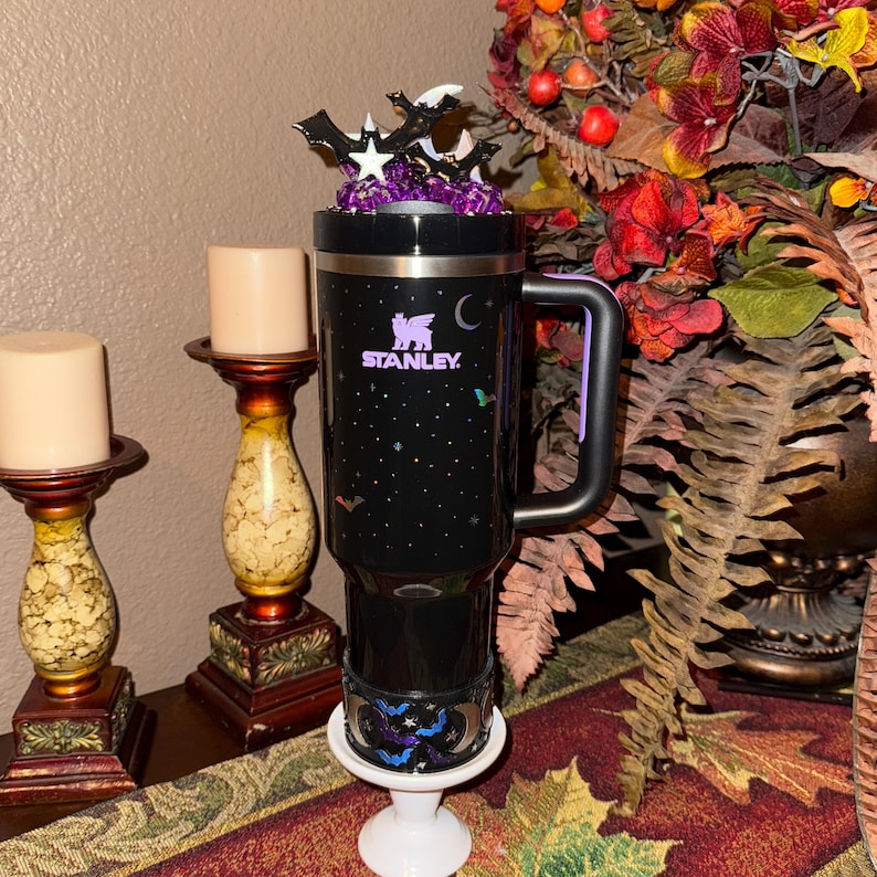 May include: Black Stanley travel mug with a handle, decorated with bats, stars, and a crescent moon. The mug has a decorative topper with bats and stars. The mug is on a white pedestal. The Stanley logo is visible.