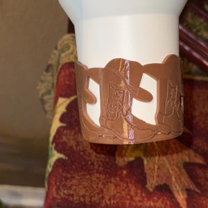 K&ouml;nnte beinhalten: Ein wei&szlig;er Becher mit einer braunen, dekorativen H&uuml;lle mit einem ausgeschnittenen Design von Cowboy-Stiefeln. Die H&uuml;lle besteht aus einem harten Material und ist um den Boden des Bechers gewickelt. Der Becher steht auf einer rot gemusterten Oberfl&auml;che.
