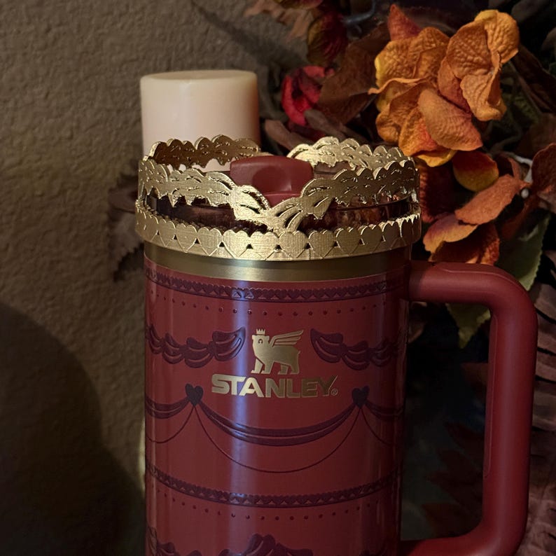 May include: A red Stanley mug with a gold-colored decorative crown lid. The mug has a handle and a gold logo. A white candle sits on top of the lid. The mug is decorated with a repeating pattern.