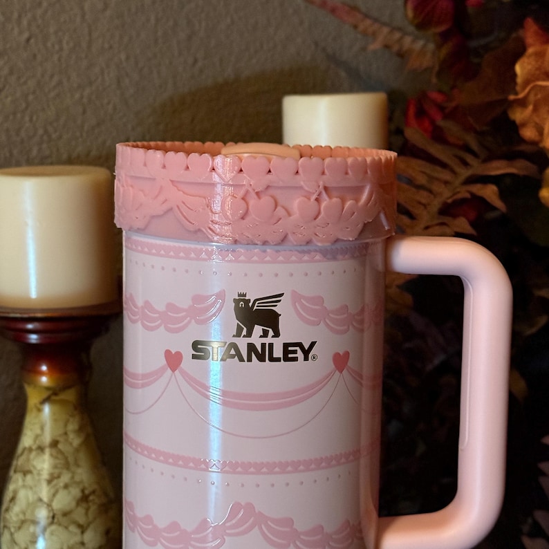 May include: Pink Stanley travel mug with a decorative floral design. The mug features the Stanley logo and a matching pink handle. The top of the mug has a decorative pink trim. The mug is designed for hot or cold beverages.
