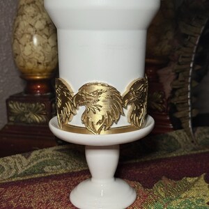 May include: A white candle holder with a gold eagle design around the middle. The eagle design is detailed and features the head and wings of the bird. The candle holder has a pedestal base and a small, round top.