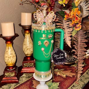 May include: A green Stanley tumbler decorated with gold and rhinestone embellishments, including a butterfly topper. The tumbler has a handle and is adorned with a variety of decorative elements, including a gold chain and a small logo.