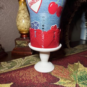 May include: A blue denim-textured tumbler featuring a red heart with the text DARLIN, red cherries, and blue dice graphics. The tumbler is held in a red, intricate, laser-cut decorative sleeve and rests on a small, white, pedestal-style stand.