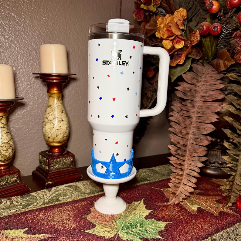 May include: White Stanley tumbler with a handle, decorated with red, blue, and black stars. The tumbler has the word "STANLEY" printed on it. It is placed on a white pedestal with a blue star-shaped design.