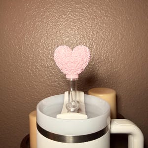 May include: A pink heart-shaped bottle stopper with a decorative floral design, attached to a clear glass tube. The stopper is placed on a white and light blue mug with a handle. The background is a textured brown wall.